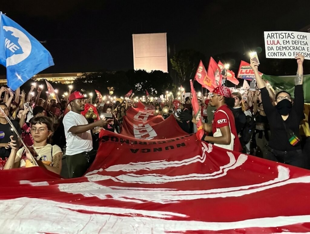 Multidão de manifestantes reunidos em frente ao Museu Nacional da República, em Brasília, segurando faixas em ato político.