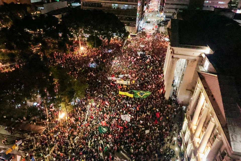 Multidão reunida durante a noite na Praça Santos Andrade, em Curitiba, com pessoas segurando bandeiras em ato político visando o segundo turno das eleições.