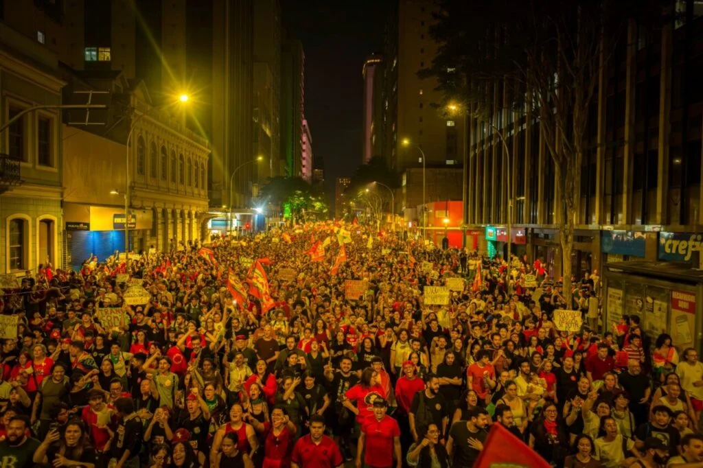 Multidão reunida durante a noite na Praça Santos Andrade, em Curitiba, com pessoas segurando bandeiras em ato político visando o segundo turno das eleições.
